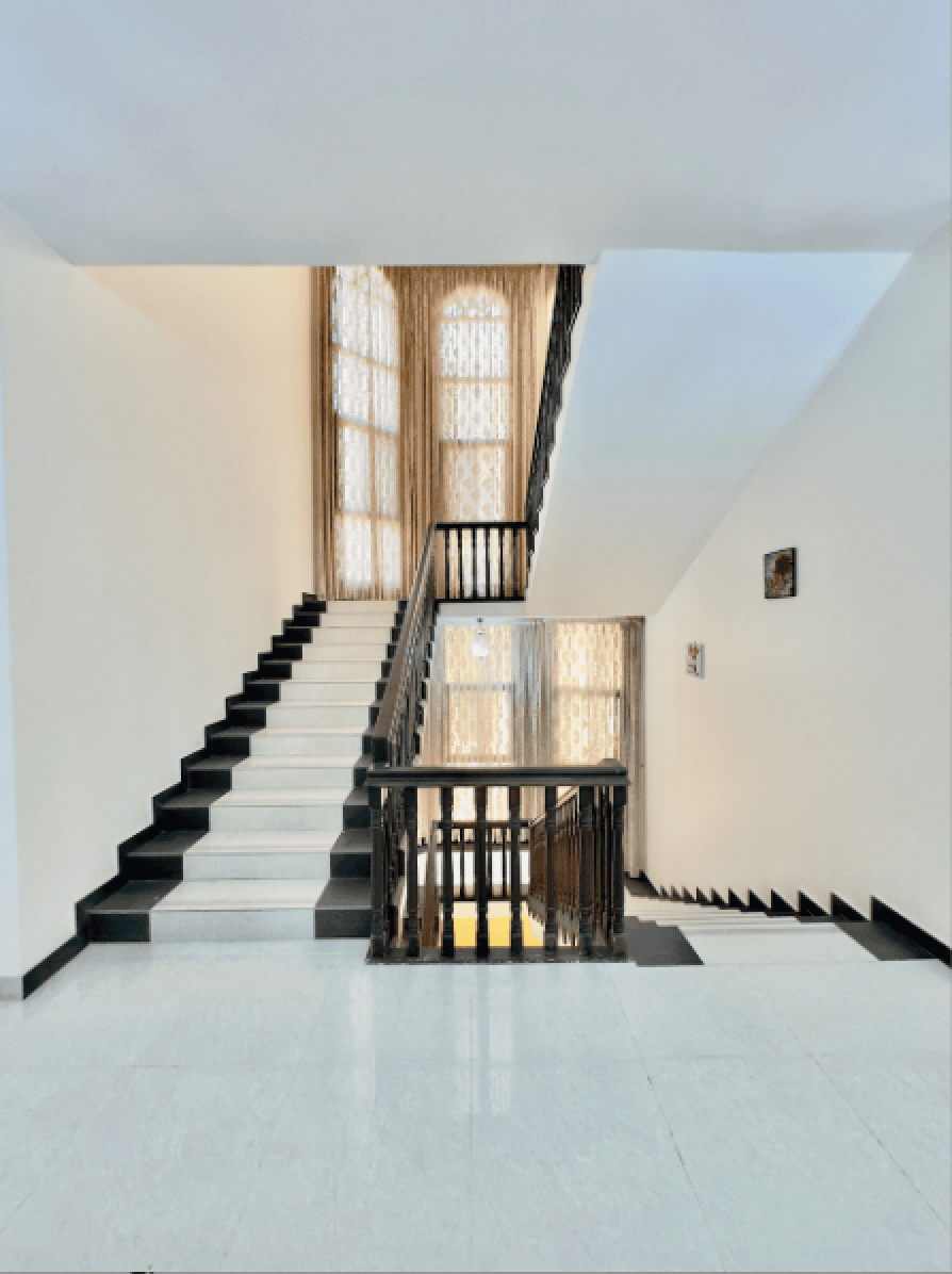 A staircase with dark wooden railings and black-and-white steps leads to upper and lower levels in a bright interior space with tall windows and white walls.